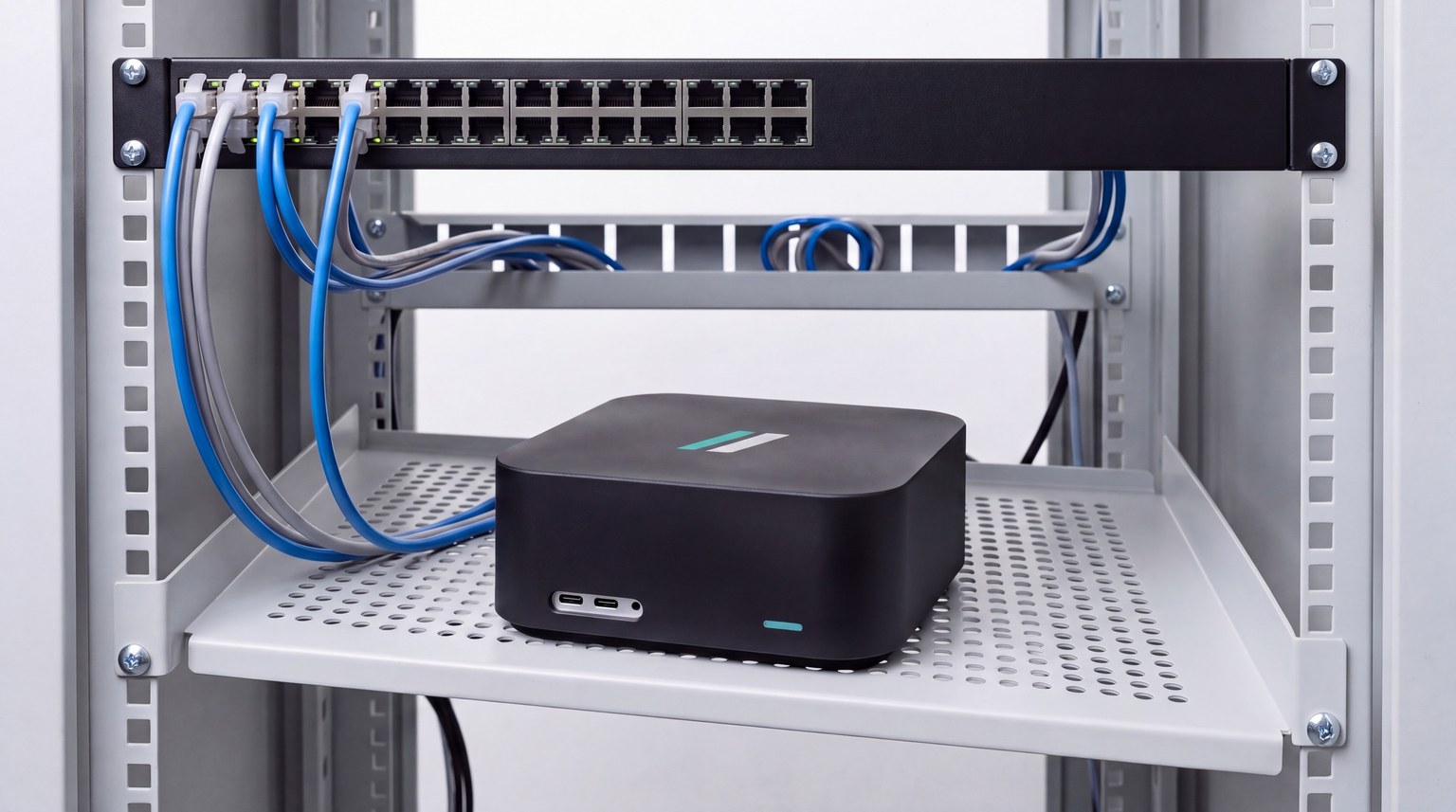 A compact satin-black razorBridge Outpost on a light-grey network cabinet shelf below a rack-mounted switch, cabled from behind, its teal status light on.